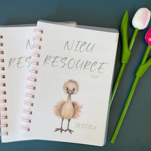 May include: A white and pink spiral bound notebook with the text "NICU RESOURCE 2024" and a drawing of a small brown bird. The notebook is on a blue surface with two green and pink flower pens.