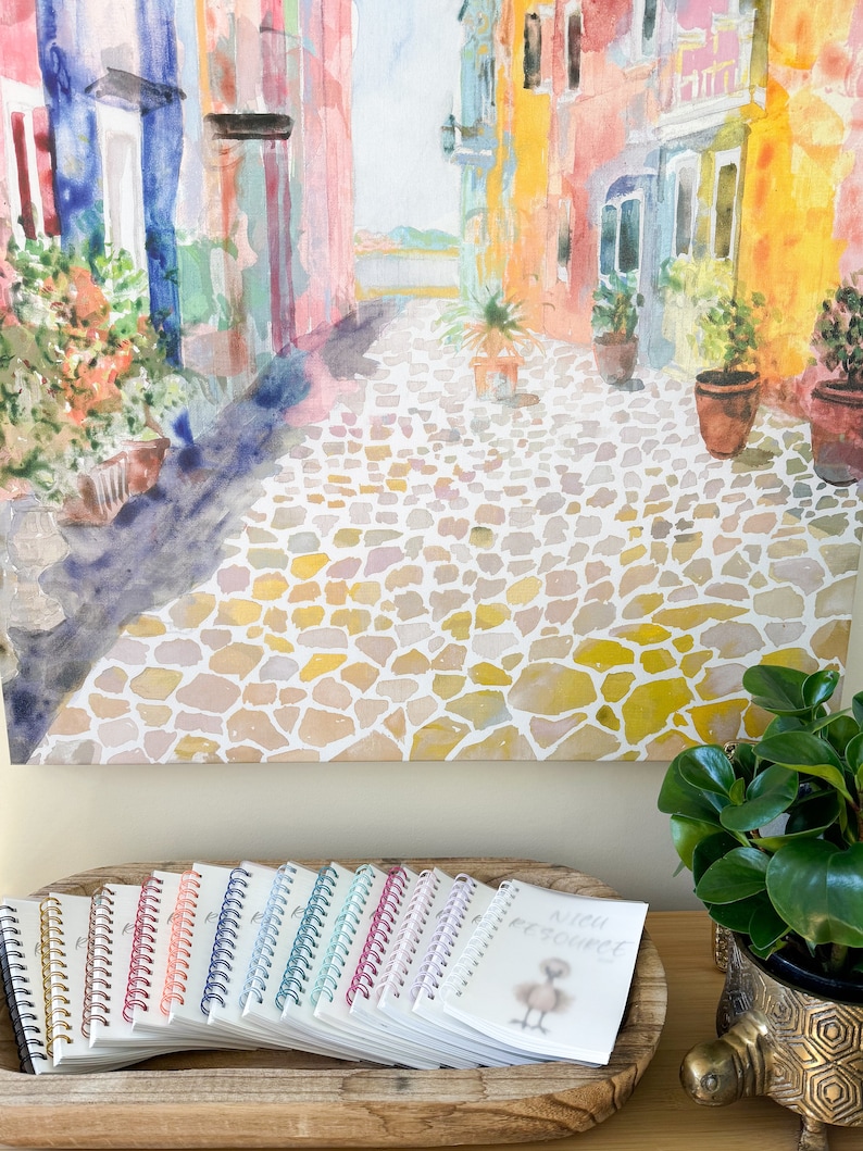 May include: A set of 12 colorful spiral bound notebooks with a watercolor painting of a cobblestone street in a European city in the background. The notebooks are arranged in a wooden tray with a small notebook with a bird illustration and the text "Nice to meet you" on the front.