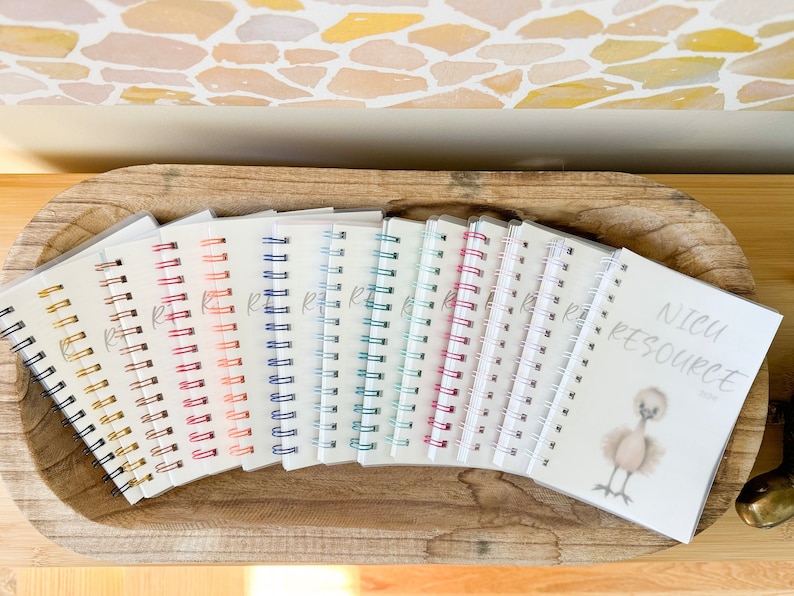 May include: A collection of 12 small spiral bound notebooks with different colored binding rings. The notebooks are arranged in a semi-circle on a wooden surface. The notebook on the right has the text "NICU RESOURCE" printed on the cover.