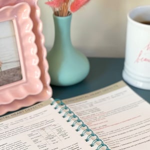 May include: A close-up of an open spiral bound book with the title "Congenital Heart Defects" on the cover. The book is on a dark blue surface with a pink picture frame, a light blue vase with pink dried flowers, and a white mug with pink lettering.