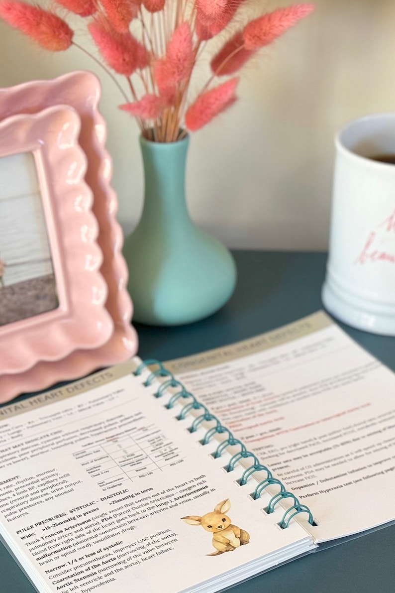 May include: A close-up of an open spiral bound book with the title "Congenital Heart Defects" on the cover. The book is on a dark blue surface with a pink picture frame, a light blue vase with pink dried flowers, and a white mug with pink lettering.