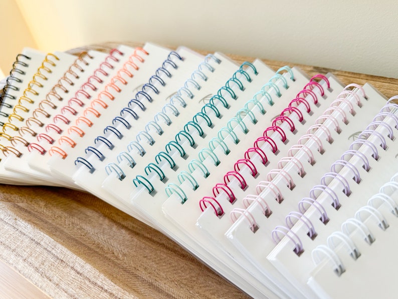 May include: A collection of spiral bound notebooks with colorful wire binding. The notebooks are arranged in a row, with the colors of the binding progressing from gold to white.
