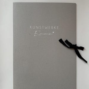 Könnte beinhalten: Ein graues Portfolio mit dem Text "KUNSTWERKE Emma" in weißer Schrift. Auf der Seite ist ein schwarzes Band gebunden. Das Portfolio besteht aus einem strukturierten Papiermaterial.