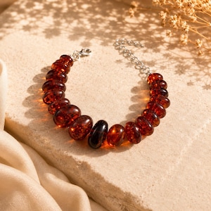 Könnte beinhalten: Ein Armband mit bernsteinfarbenen, oval geformten Perlen, die mit einer silbernen Kette und einem Verschluss aufgereiht sind. Die Perlen variieren in der Farbe von hell bis dunkel Bernstein, mit einer schwarzen Perle. Das Armband liegt auf einer neutralen Oberfläche.