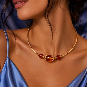 Op de afbeelding: Een goudkleurige choker ketting met een grote, barnsteenkleurige kraal in het midden, geflankeerd door twee kleinere barnsteenkleurige kralen. De kralen zijn voorzien van kleine goudkleurige metalen tussenstukjes op een flexibele draad.