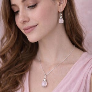 Op de afbeelding: Een zilveren ketting en bijpassende oorbellen set met lichtroze, gefacetteerde edelsteen hangers. De sieraden worden gepresenteerd tegen een zachte roze achtergrond, wat een delicate esthetiek creëert. De ketting hangt aan een zilveren ketting.