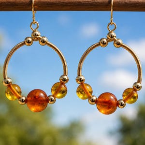 May include: A pair of gold-toned hoop earrings featuring a coiled wire design accented with polished gold-toned beads and translucent, round amber-colored beads. The earrings hang from gold-toned hooks and display a symmetrical arrangement of beads.