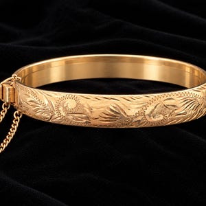 Puede incluir: Un brazalete rígido articulado de tono dorado con un intrincado patrón floral grabado en la superficie exterior. La pulsera incluye un mecanismo de cierre seguro y una delicada cadena de seguridad adjunta, con un diseño circular.