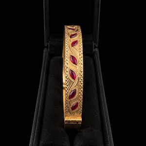 Può includere: Un bracciale rigido incernierato color oro con una serie di pietre rosso scuro a taglio marquise disposte in fila. La superficie metallica è decorata con un intricato motivo inciso a tratteggio incrociato e volute lungo tutta la fascia.