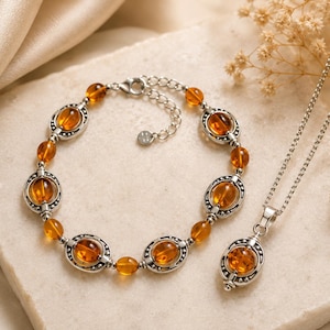 Può includere: Un set di bracciale e collana in argento con pietre ovali color ambra. Il bracciale e la collana presentano pietre ovali color ambra incastonate in argento. I gioielli sono esposti su una superficie neutra.