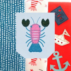 Può includere: Una toppa ricamata azzurra con un'aragosta rosa con zampe e coda blu e chele verdi. L'immagine è circondata da tessuti con motivi nautici, tra cui aragoste rosse e bianche e un'ancora blu.