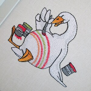Op de afbeelding: Borduurwerk van een witte gans die zichzelf schildert met een kwast. De gans houdt een kwast in zijn vleugel en heeft verfspatten op zijn lichaam. Er zijn drie verfpotten naast de gans.
