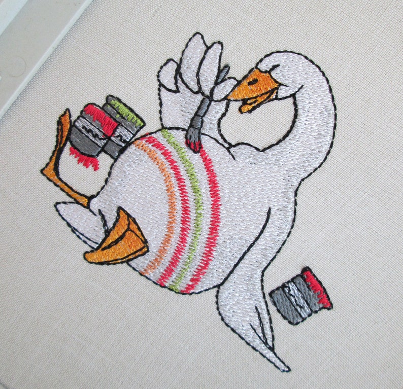 Op de afbeelding: Borduurwerk van een witte gans die zichzelf schildert met een kwast. De gans houdt een kwast in zijn vleugel en heeft verfspatten op zijn lichaam. Er zijn drie verfpotten naast de gans.