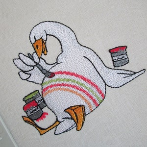 Op de afbeelding: Borduurwerk van een witte gans die een kwast vasthoudt en naast twee verfpotten staat. De gans heeft een rood-oranje-groen gestreept patroon op zijn lichaam.