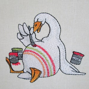 Op de afbeelding: Borduurwerk van een witte gans die zichzelf schildert met een kwast. De gans zit op zijn zij en heeft een regenboog van kleuren op zijn rug geschilderd. Er staan twee verfpotten naast de gans.
