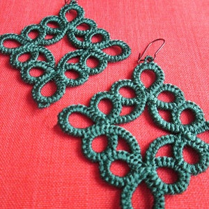 Puede incluir: Un par de pendientes de ganchillo verde con un diseño delicado e intrincado. Los pendientes son de forma cuadrada y presentan un patrón repetitivo de bucles y nudos.