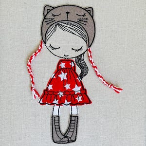 May include: Embroidered artwork of a girl wearing a cat-eared hat and a red dress with white stars. The girl has long black hair and is wearing gray boots. Braided red and white yarn hangs from the hat.