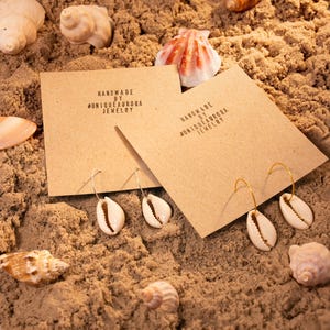 Könnte beinhalten: Zwei Paar handgefertigte Ohrringe mit Kaurimuscheln, präsentiert auf Sand mit Muscheln. Die Ohrringe befinden sich auf braunen Karten mit dem Text "HANDMADE BY @UNIQUEAURORA JEWELRY". Ein Paar hat silberne, das andere goldene Creolen.