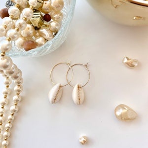 Puede incluir: Un par de pendientes de aro dorados con colgantes de concha blanca. Los pendientes están sobre una superficie blanca con un collar de perlas y otras cuentas en el fondo.