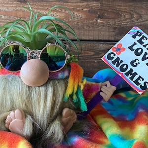 May include: A gnome wearing tie-dye clothing and sunglasses holds a sign that says "Peace, Love & Gnomes". The gnome is sitting on a colorful tie-dye blanket.