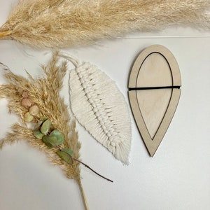 Könnte beinhalten: Eine weiße Makramee-Feder hängt neben einem hölzernen, blattartigen Rahmen mit einem schwarzen Gummiband. Die Gegenstände befinden sich auf einer weißen Oberfläche mit getrocknetem Pampasgras.