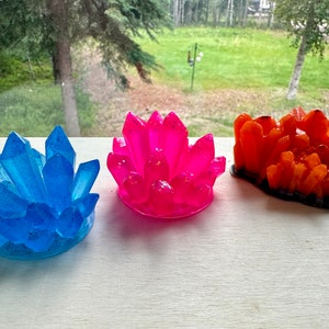 Op de afbeelding: Drie kristalvormige decoratieve objecten in blauw, roze en oranje. Het blauwe kristal heeft glitteraccenten. Het roze kristal staat in het midden en het oranje kristal rechts. Alle drie staan op een wit oppervlak.