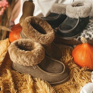 Può includere: Due paia di pantofole in camoscio marrone con un bordo in pelliccia sintetica marrone soffice. Le pantofole sono appoggiate su una superficie marrone soffice con una zucca sullo sfondo.