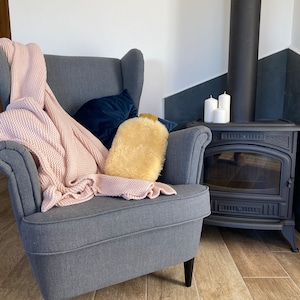 May include: A gray armchair with a pink knitted throw blanket and a fluffy white cushion. The chair is in front of a black wood-burning stove with white candles on top.