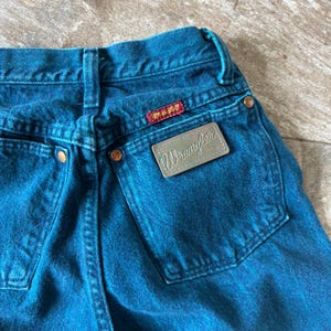 May include: Close-up of blue denim jeans with the Wrangler brand label on the back pocket. The jeans have copper-colored rivets and a red and yellow brand tag. The denim is a solid, medium blue color.