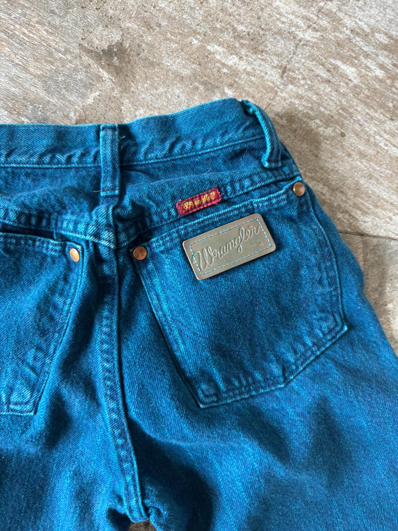 May include: Close-up of blue denim jeans with the Wrangler brand label on the back pocket. The jeans have copper-colored rivets and a red and yellow brand tag. The denim is a solid, medium blue color.