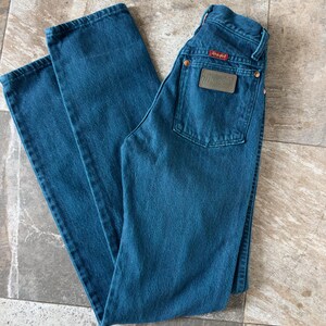 May include: A pair of blue denim jeans with a classic five-pocket design. The jeans feature a leather patch on the back pocket with the brand name "Wrangler". The jeans are laid flat on a tiled surface.