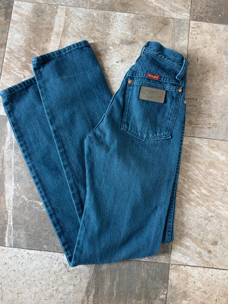 May include: A pair of blue denim jeans with a classic five-pocket design. The jeans feature a leather patch on the back pocket with the brand name "Wrangler". The jeans are laid flat on a tiled surface.