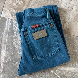May include: Folded pair of teal Wrangler jeans. The jeans feature a brown leather patch with the Wrangler logo and a red tag. The jeans have a classic five-pocket design with copper-colored rivets and a button closure.