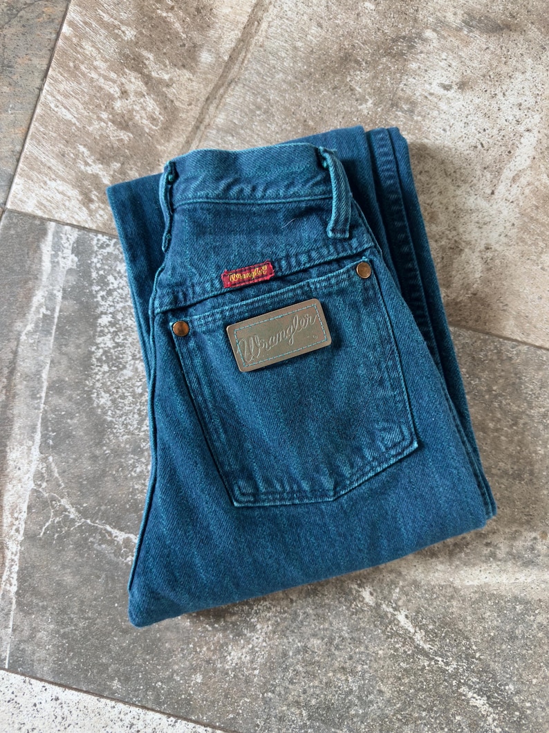 May include: Folded pair of teal Wrangler jeans. The jeans feature a brown leather patch with the Wrangler logo and a red tag. The jeans have a classic five-pocket design with copper-colored rivets and a button closure.