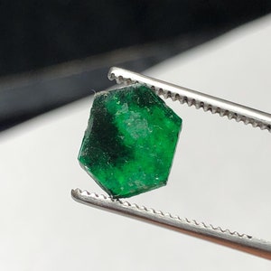 Könnte beinhalten: Ein leuchtend grüner Smaragd, der mit einer Pinzette gehalten wird. Der Smaragd hat einen facettierten Schliff und ist von einer tiefen, satten grünen Farbe.