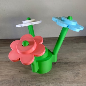Pode incluir: Um vaso verde com três flores. As flores são rosa, branca e azul claro. As flores são feitas de plástico e têm um design simples.