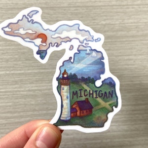 Può includere: Un adesivo che rappresenta lo stato del Michigan con un faro, una piccola casa e la scritta "MICHIGAN" nell'angolo in basso a destra. L'adesivo è un'illustrazione in stile acquerello con una gamma di colori blu e verdi.