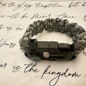 May include: Gray paracord survival bracelet with a black clasp and a small compass. The bracelet is made of woven gray and white cord. The clasp is black and has a built-in compass. The background is a white paper with handwritten text.