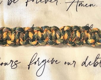 Può includere: Un braccialetto in paracord intrecciato nei toni arancione, nero e verde, con chiusura a fibbia in plastica nera. Il braccialetto è appoggiato su una superficie color crema con testo scritto. Lunghezza bracciale: circa 20 cm.
