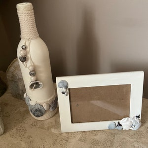 May include: A white painted wine bottle decorated with seashells and rope, and a white picture frame with seashells on the edges. Both items are on a beige surface.