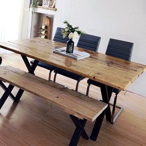 Puede incluir: Una mesa de comedor de madera rústica con patas de metal negro y un banco de madera a juego. La mesa está puesta con un jarrón de flores blancas y una bandeja con posavasos.