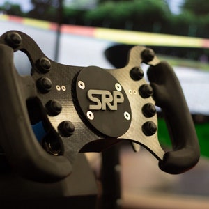 Op de afbeelding: Close-up van een zwart race stuur met een carbon fiber afwerking. Het stuur heeft meerdere knoppen en een ronde, zwarte middenplaat met het woord "SRP" in zilveren letters.