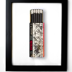 May include: A black framed picture of a matchbox with a white background. The matchbox is red and white with the text "Espolón Tequila Blanco Mexico" and a black and white illustration of skeletons dancing. The matchbox is open and there are six matches inside.