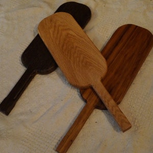 May include: Three wooden serving paddles, one dark brown, one light brown, and one medium brown, with handles. The paddles are arranged in a criss-cross pattern on a white surface.