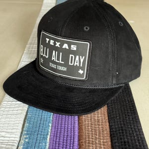 Könnte beinhalten: Schwarze Baseballkappe mit einem weißen Patch, auf dem "TEXAS BJJ ALL DAY TEXAS TOUGH" steht. Die Kappe liegt auf einem Stapel bunter Gürtel.