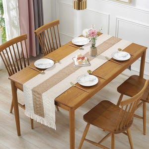 May include: A wooden dining table set for a meal, with a beige and brown table runner with fringe. The table is set with white plates, gold utensils, and a vase of flowers. Wooden chairs surround the table, creating a warm and inviting atmosphere.