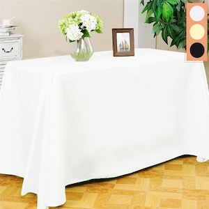 May include: White rectangular tablecloth draped over a table with a vase of white flowers and a framed picture on top. The table is on a wooden floor.
