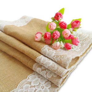May include: A burlap table runner with a white lace trim. The runner is folded in thirds and decorated with a bouquet of pink and red flowers.