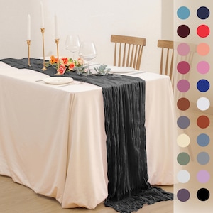 May include: A black linen table runner on a white tablecloth with gold candlesticks and flowers. The table runner is draped over the table and has a textured look. Two chairs are visible in the background.