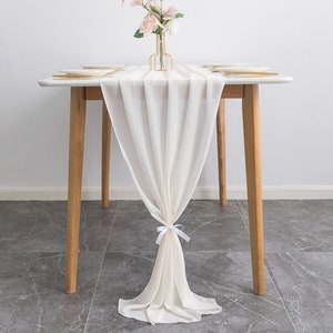 May include: A white chiffon table runner with a white bow tied at the centre. The runner is draped over a wooden table with a vase of pink flowers in the centre.